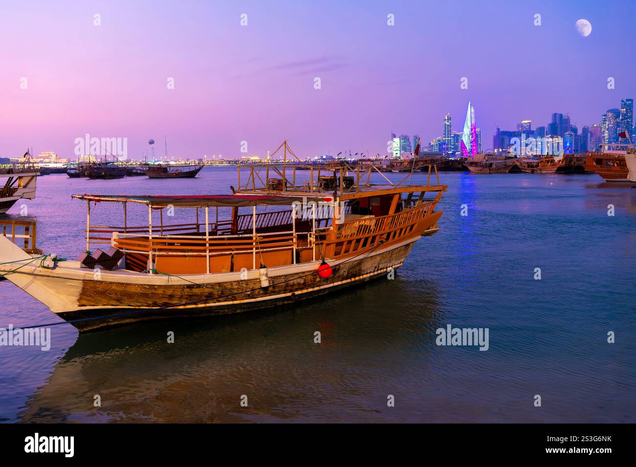 Barche tradizionali in dhow lungo la Corniche sul lungomare con lo skyline illuminato in lontananza all'ora blu crepuscolo, a Doha in Qatar. Foto Stock