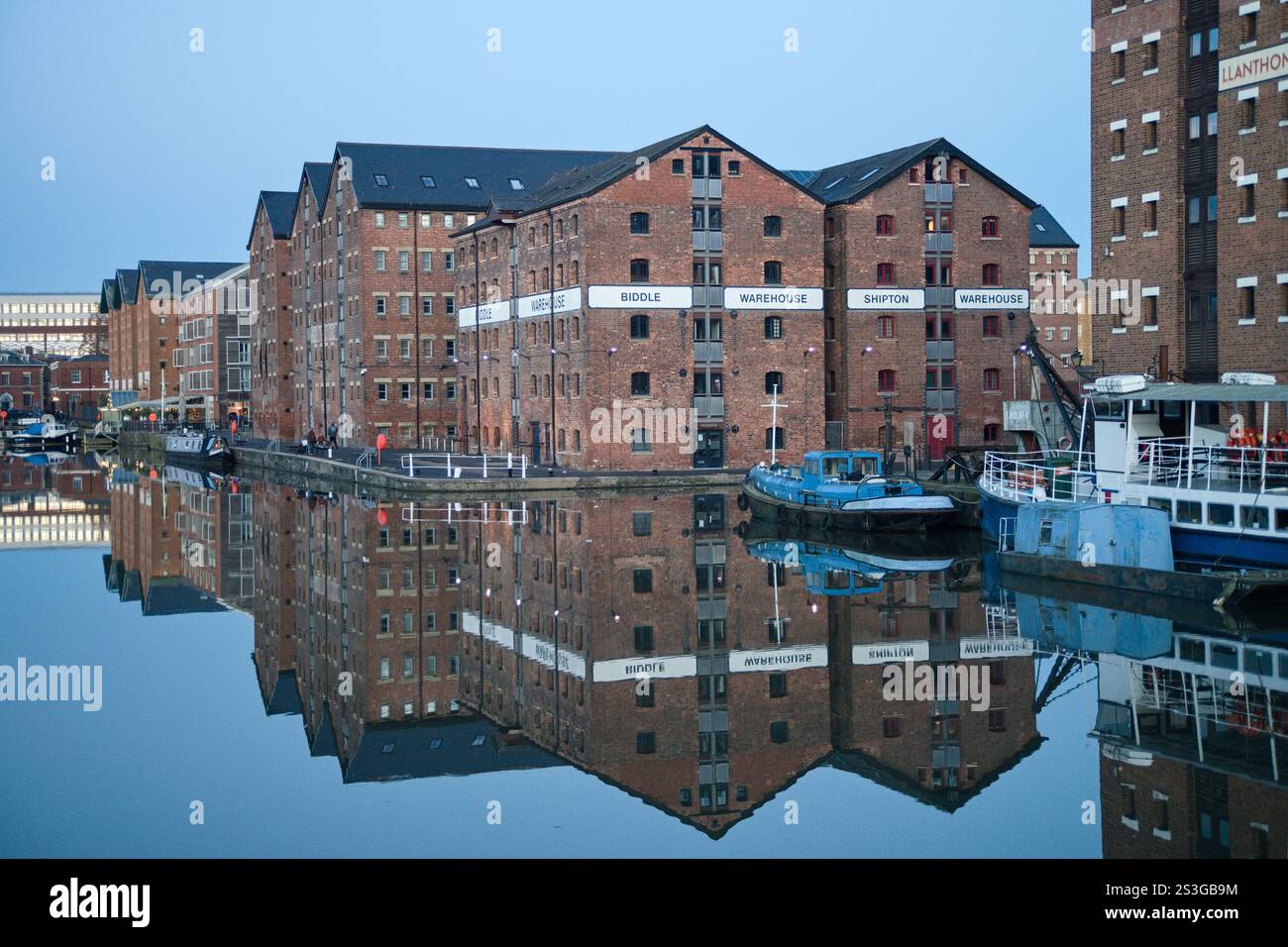 Una fila di storici magazzini in mattoni che si riflettono nelle calme acque di Gloucester Quays al crepuscolo. Gli edifici hanno una grandiosità sbiadita e creano un senso di Foto Stock