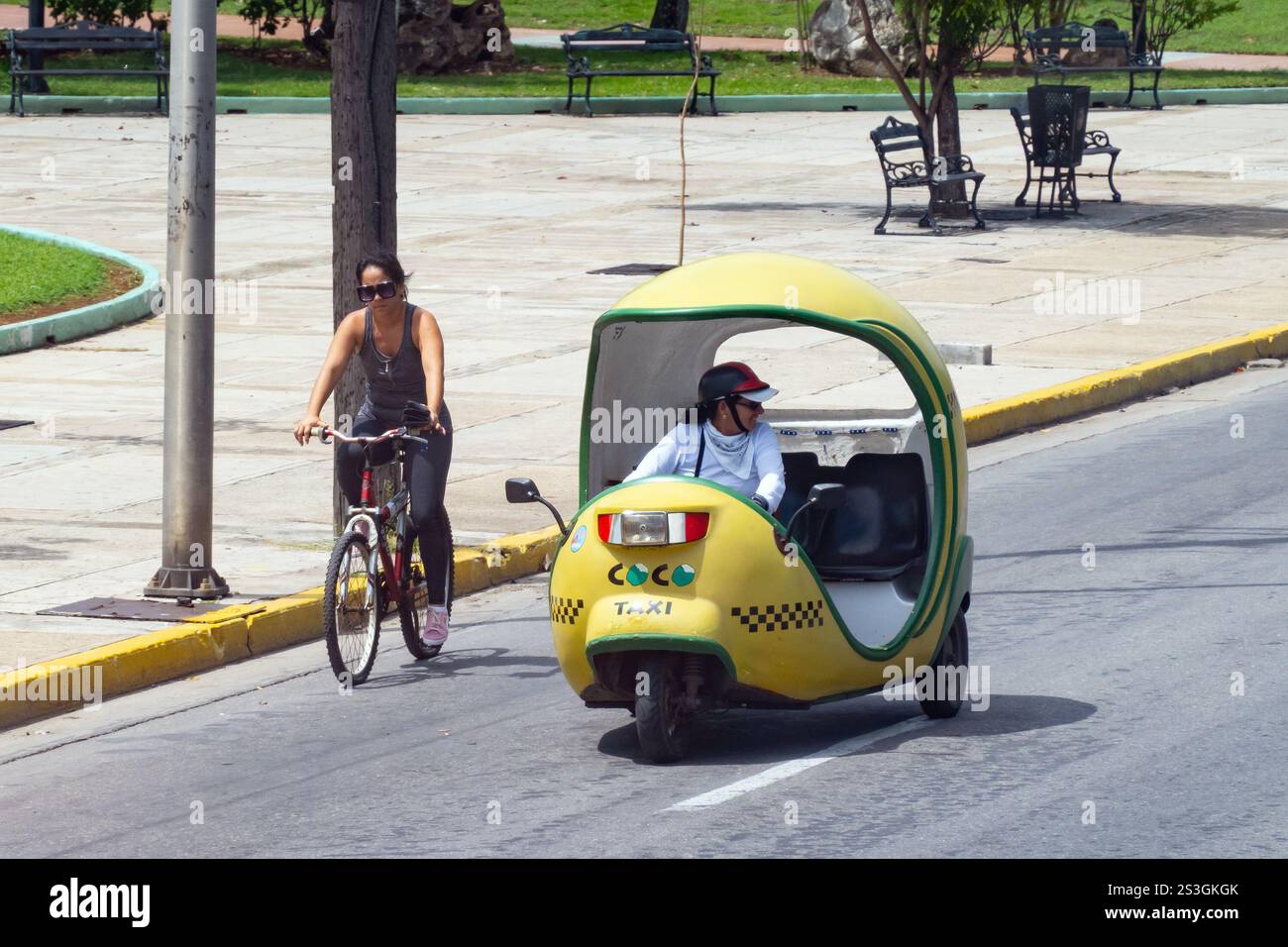 VARADERO, CUBA - 30 AGOSTO 2023: Rickshaw giallo tuk-tuk veicolo coco-taxi nelle strade di Varadero, Cuba e donna in bicicletta Foto Stock