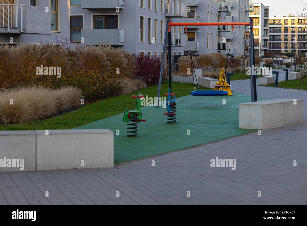 Moderno parco giochi con altalene colorate e ciclisti primaverili su una superficie sicura in gomma circondata da erbe decorative ed edifici residenziali Foto Stock
