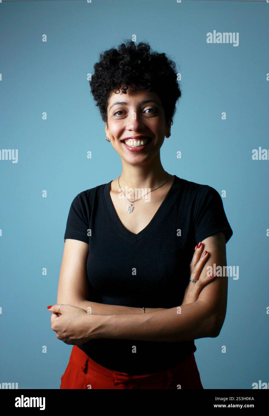 Ritratto verticale di donna brasiliana in pelle marrone, con t-shirt nera e collana in argento, di fronte alla parete blu. Colpo di studio, allegra signora. Foto Stock