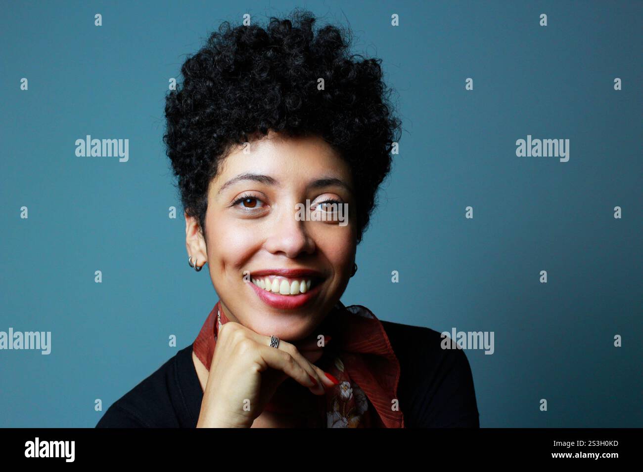 Donna dalla pelle marrone che sorride e guarda la fotocamera. Modello femminile con capelli ricci corti. Donna afro americana che indossa camicia nera e sciarpa marrone. Foto Stock