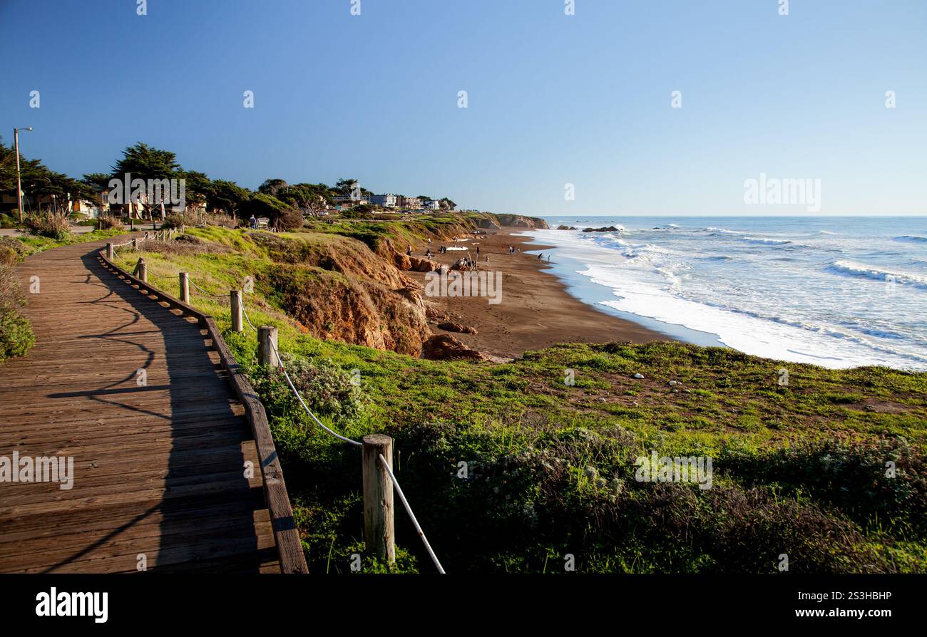 Una passerella in legno conduce lungo la spiaggia di Moonstone, caratterizzata da dolci onde e lussureggiante vegetazione sotto un cielo limpido, che mostra la bellezza dei cosi' come della California Foto Stock