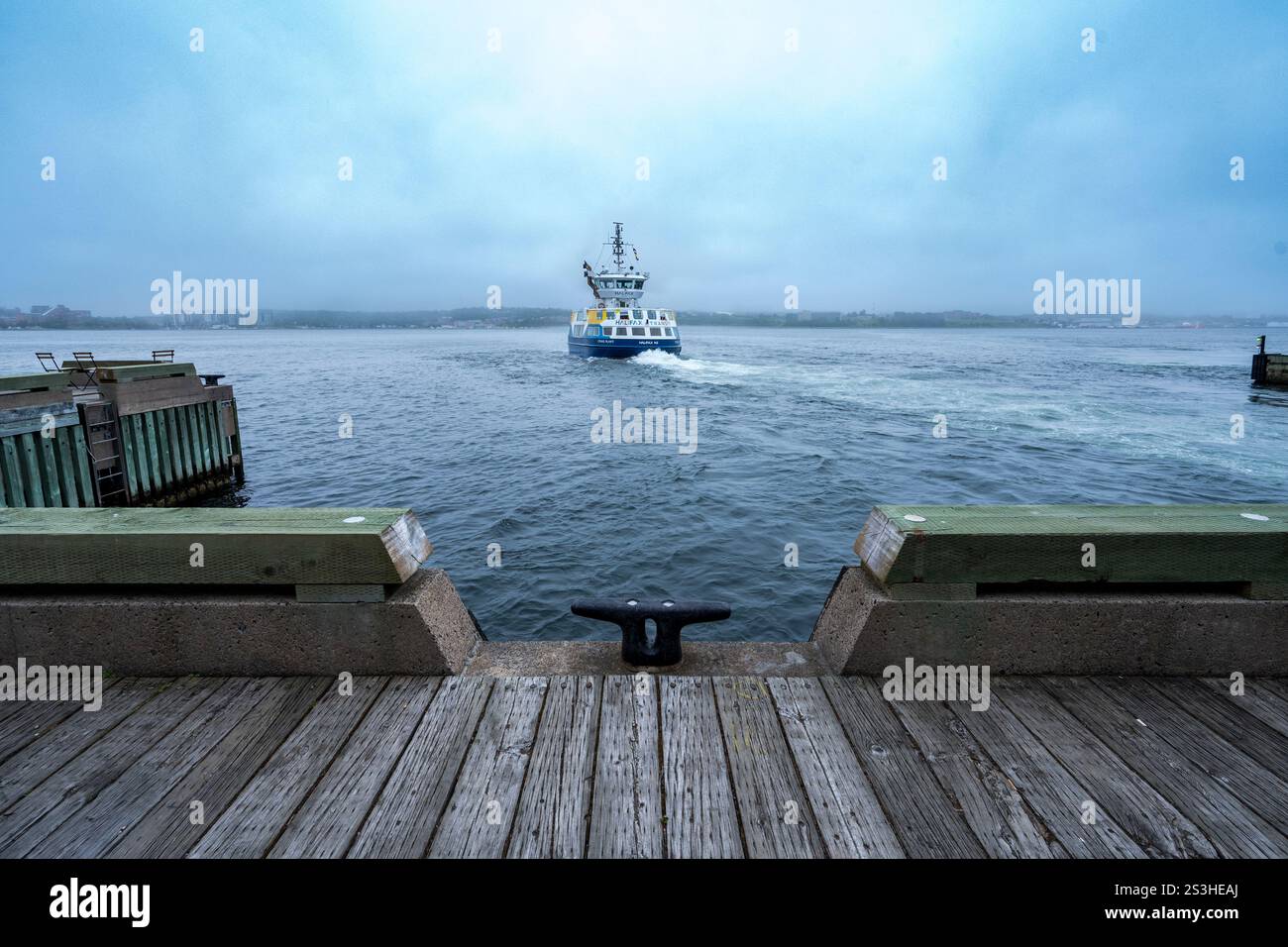Il traghetto Halifax Transit parte per Dartmouth, catturato contro uno skyline nebbioso. La sua scia si snoda dietro, con il molo che incornicia la tranquilla scena del lungomare. Foto Stock