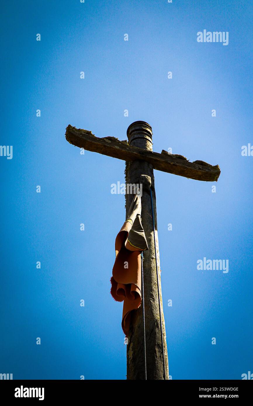 Vista ad angolo basso di una rustica croce di legno contro un cielo azzurro. Le texture e i resti di tessuto avvolti intorno al palo simboleggiano la fede, resili Foto Stock