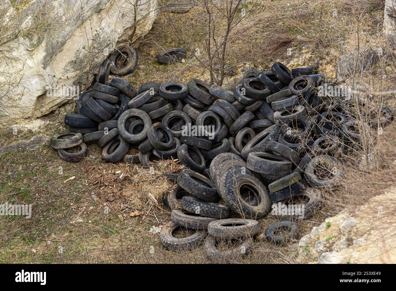 Un grande mucchio di pneumatici usurati poggia su uno sfondo roccioso, circondato da erba secca e alberi sparsi, indicando un sito di scarico illegale in un rur Foto Stock