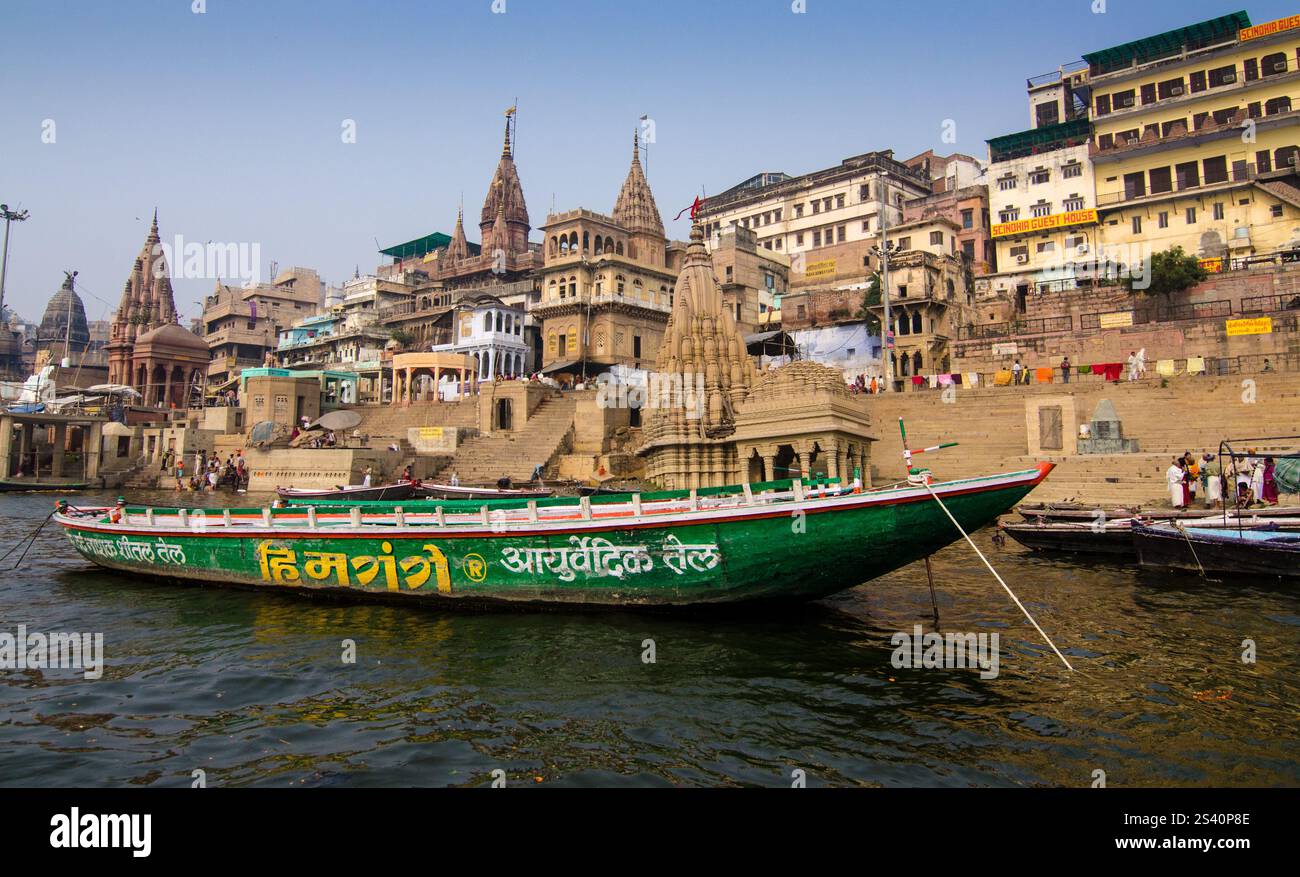 Vista dell'architettura storica lungo la riva del fiume con barche colorate a Varanasi durante il giorno Foto Stock