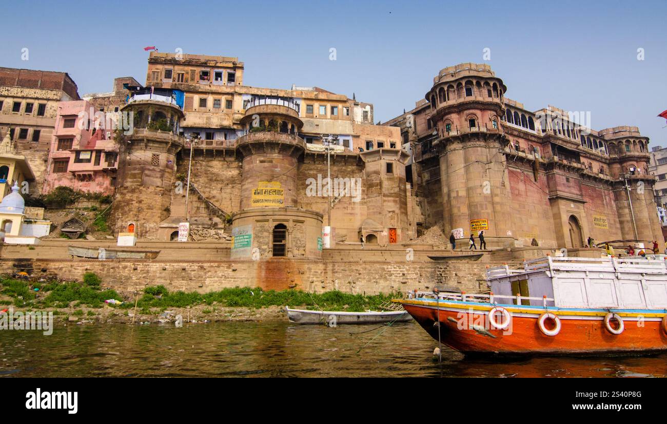Vista dell'architettura storica lungo la riva del fiume con barche colorate a Varanasi durante il giorno Foto Stock