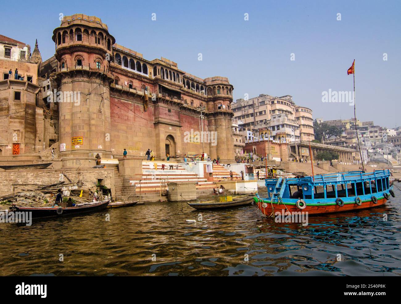 Vista dell'architettura storica lungo la riva del fiume con barche colorate a Varanasi durante il giorno Foto Stock