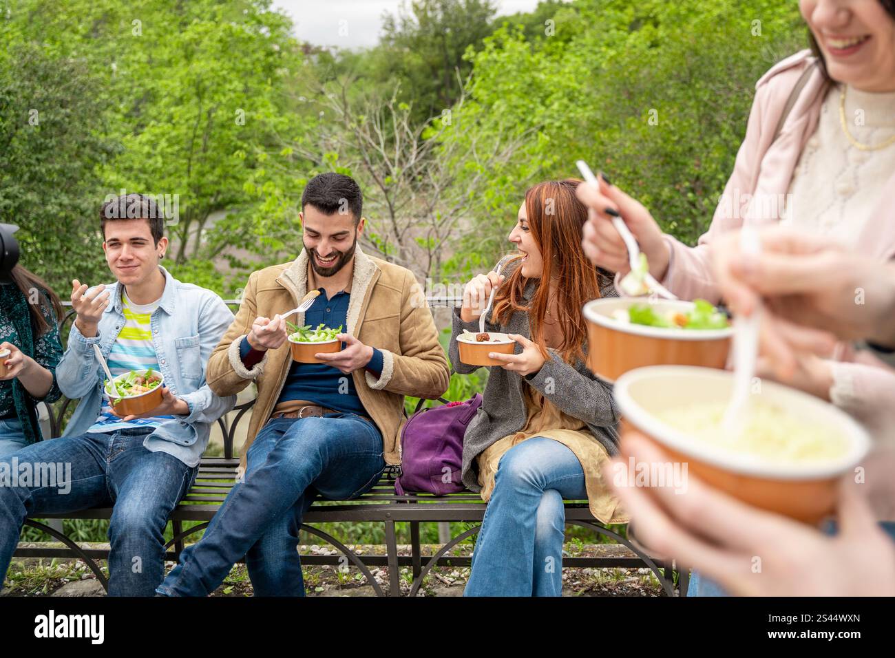 Gruppo di giovani amici seduti su una panchina in un parco, gustando pasti da asporto in ciotole ecologiche e prive di plastica. Atmosfera allegra, cornice verde Foto Stock