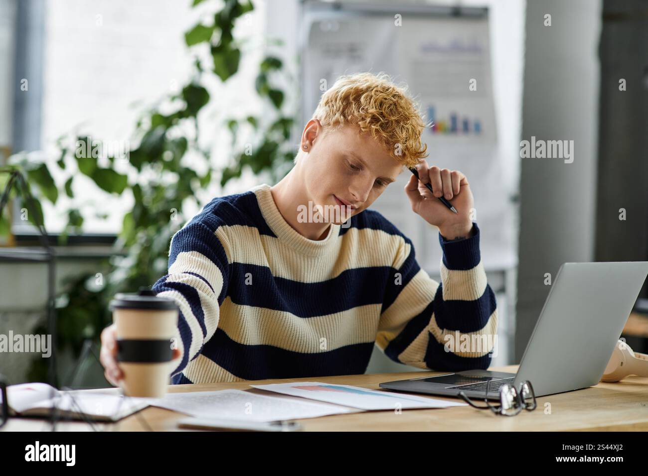 Un giovane rosso si sta concentrando sul suo lavoro in un ufficio contemporaneo mentre si gusta un caffè. Foto Stock