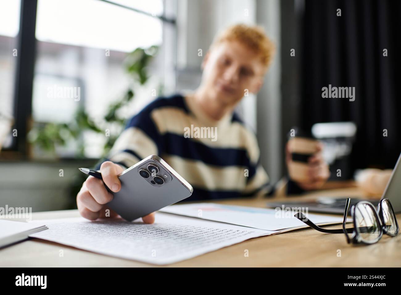 Il giovane con un maglione a righe esamina i documenti mentre utilizza uno smartphone in ufficio. Foto Stock