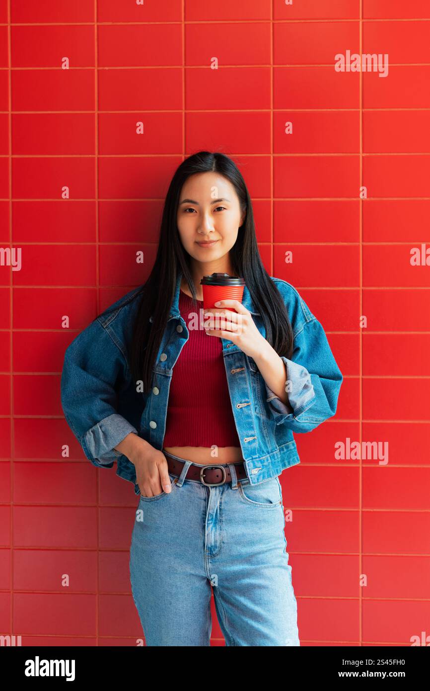 Ritratti la giovane donna asiatica coreana di generazione Z in abbigliamento casual usando una tazza di caffè di carta usa e getta che sembra felice, in piedi su una parete di mattoni rossi Foto Stock