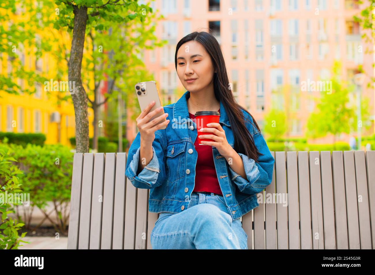 Donna asiatica coreana che usa uno smartphone e siede su una panchina nel giardino pubblico della città, una tazza di caffè usa e getta Foto Stock