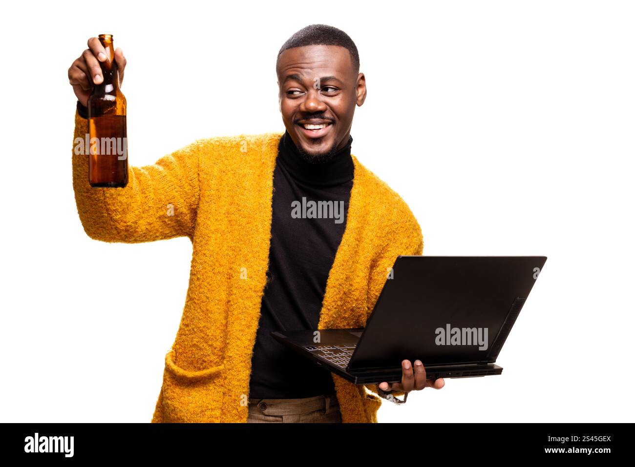 Un uomo allegro che indossa un maglione regge una bottiglia di birra e un laptop, incarnando un mix di lavoro e relax. Foto Stock