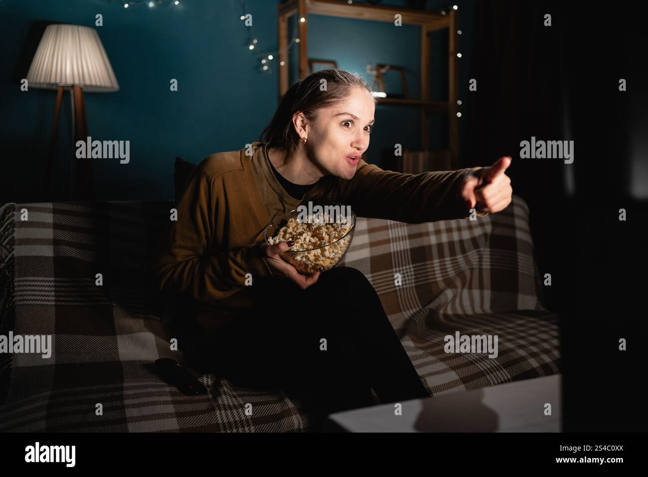 Donna emotiva che mangia mais guardando un film in TV commentando la scena sorridendo e puntando il dito sullo schermo Foto Stock