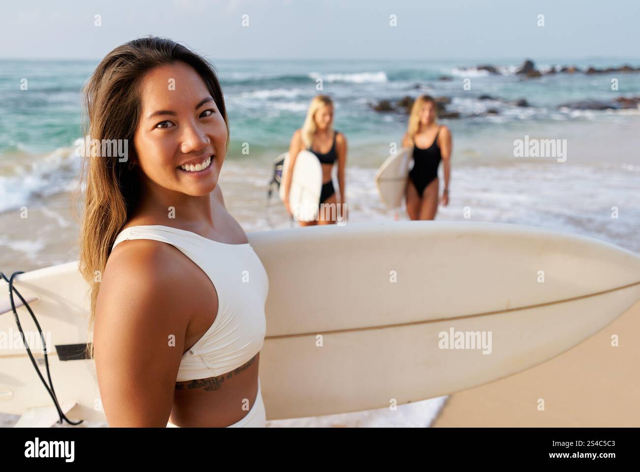 Donna asiatica con tavola da surf sorride sulla spiaggia. Due donne surfiste in sottofondo a onde oceaniche. Surf camp estivo per principianti. Spiaggia tropicale attiva Foto Stock