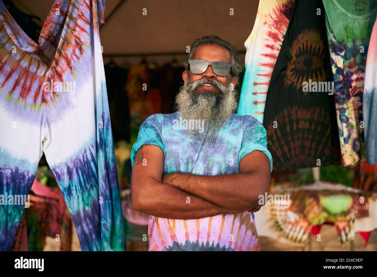 Uomo anziano con la barba lunga che indossa una t-shirt tie-dye alla fiera dell'artigianato all'aperto. Sorridenti, braccia incrociate, circondate da vestiti colorati. Azioni di artisti anziani Foto Stock