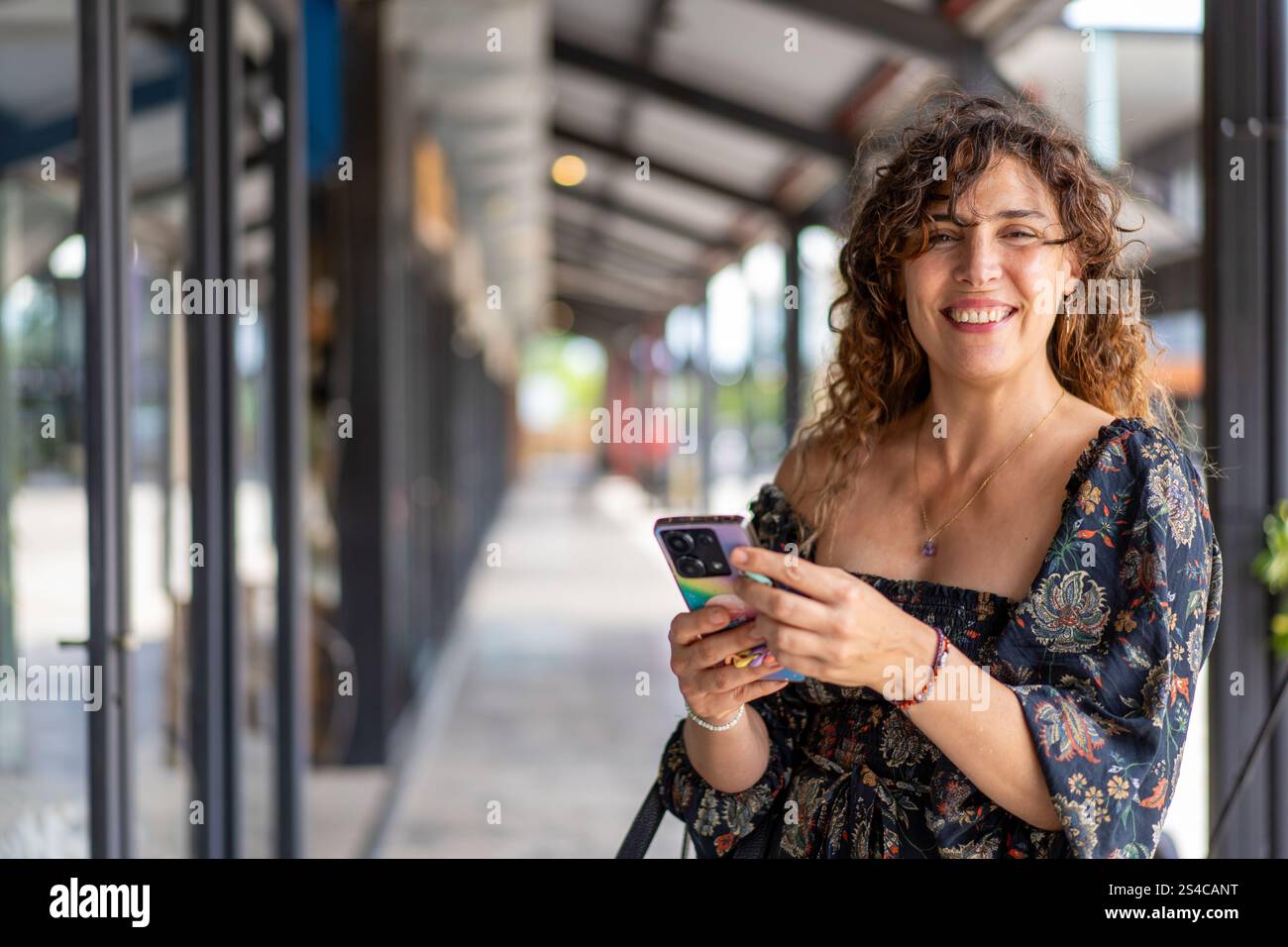Donna riccia di mezza età che usa il suo smartphone mentre guarda la fotocamera in un moderno quartiere dello shopping Foto Stock