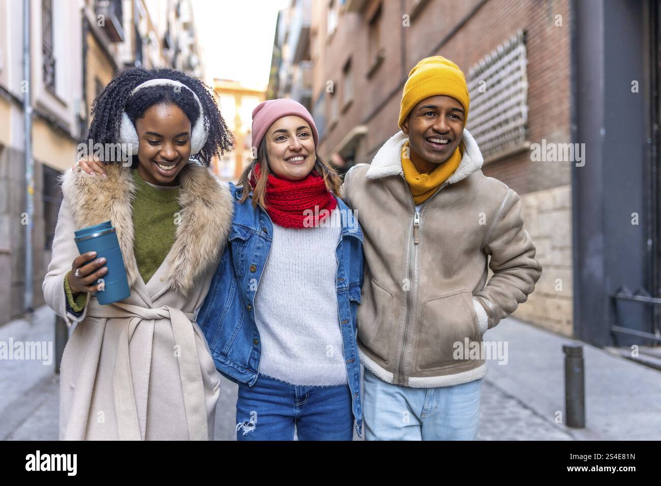 Amici multietnici felici si godono una giornata invernale insieme, camminando a braccia aperte attraverso una strada della città Foto Stock