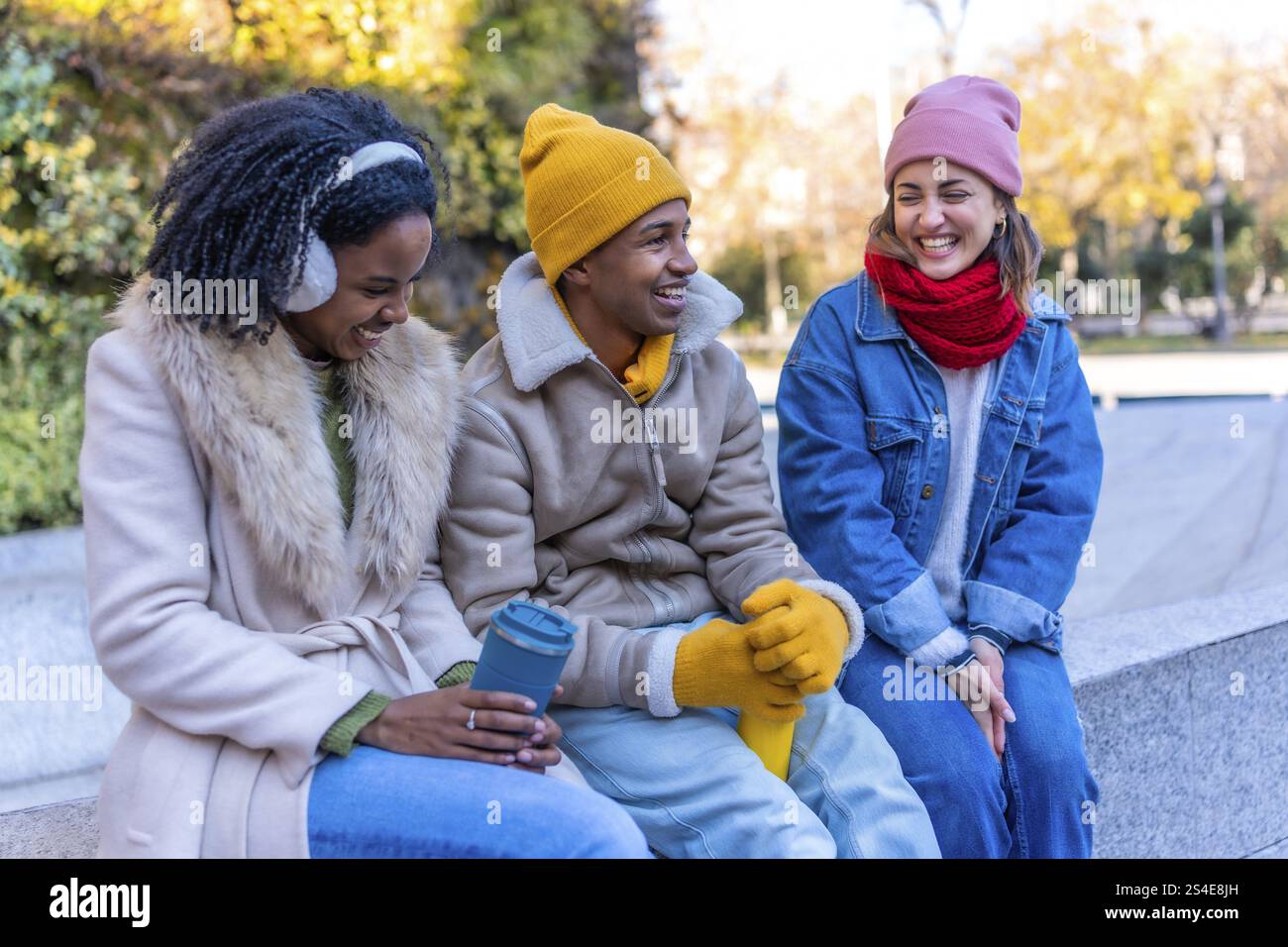 Tre giovani amici multietnici che indossano abiti invernali chiacchierando e ridendo, seduti su una panchina in un parco cittadino, durante una giornata invernale di sole Foto Stock
