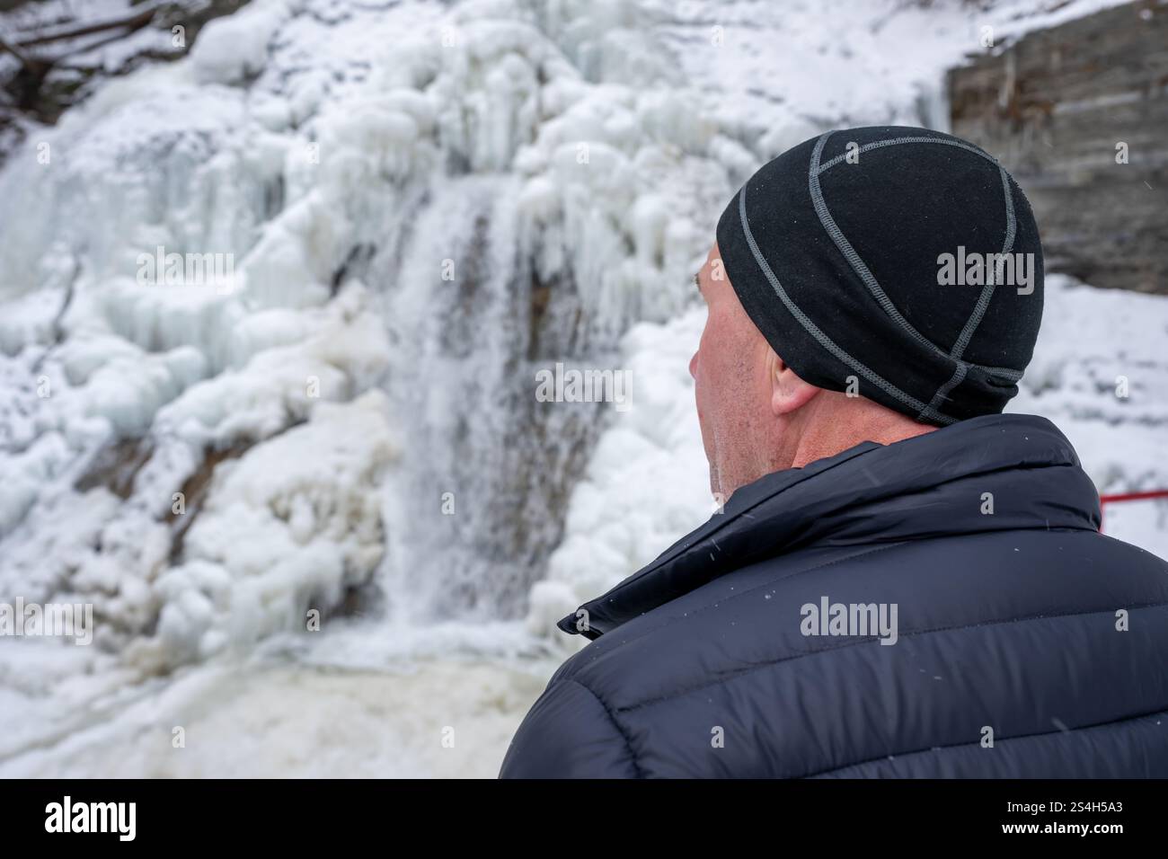 Scena invernale di un uomo con giacca nera e cappello nero che guarda una cascata ghiacciata. Prendersi del tempo per la riflessione personale e l'introspezione. Foto Stock