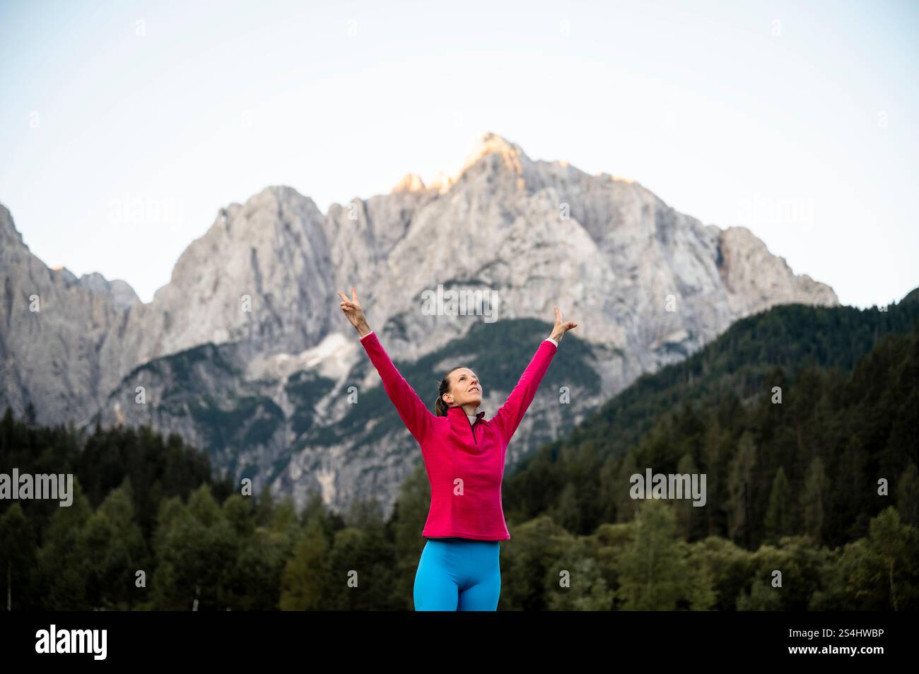 Una donna con giacca rosa e leggings blu alza con gioia le braccia, in piedi su uno sfondo mozzafiato di montagne aspre e foreste lussureggianti. Foto Stock