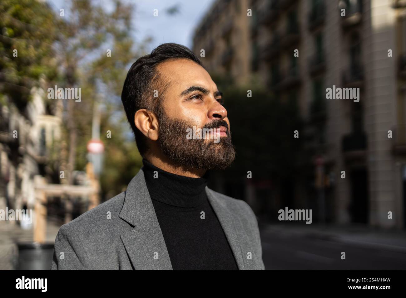 Un fiducioso uomo indiano vestito con un cappotto grigio adatto agli affari e dolcevita nero viene catturato guardando verso l'alto su una strada illuminata dal sole della città, che incarna Foto Stock