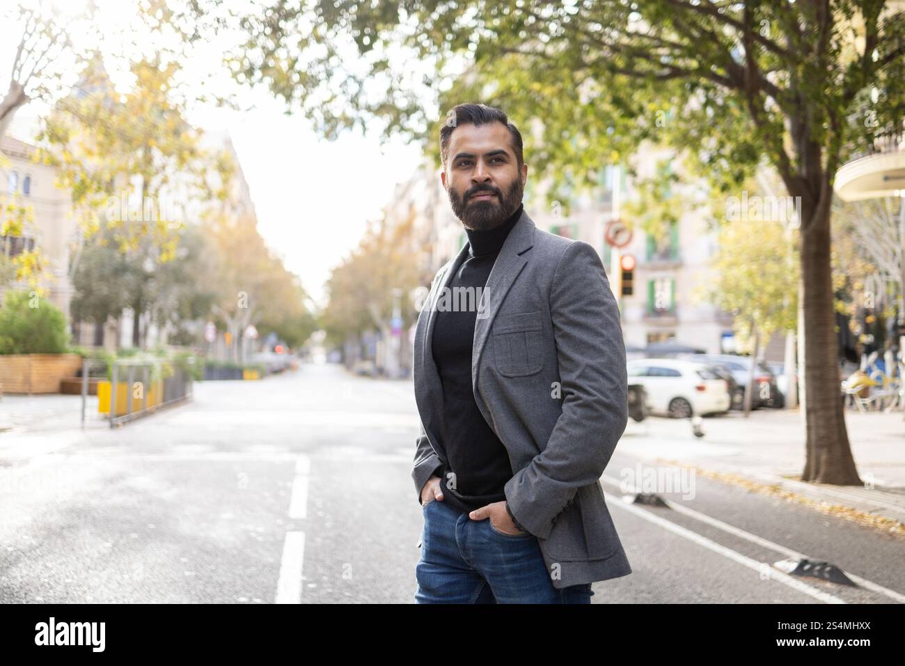 L'elegante uomo d'affari indiano con una barba ben rifinita si trova in una strada urbana costeggiata da alberi, vestito da un blazer grigio affilato e scuro Foto Stock