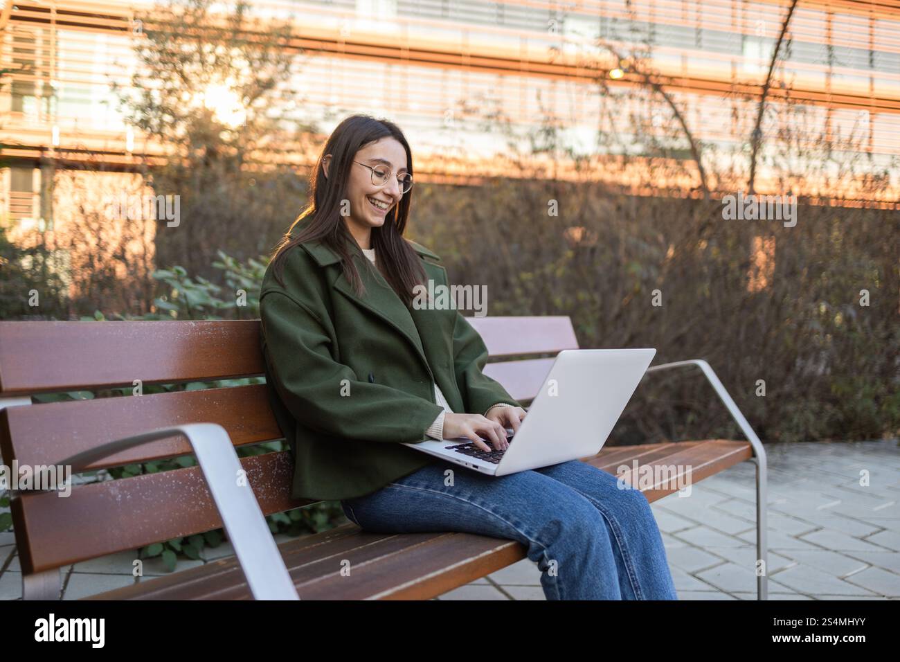 Una donna con una giacca verde e un jeans blu siede su una panchina di legno del parco, sorridendo mentre digita sul suo laptop il sole tramonta getta una calda luce sull'urba Foto Stock