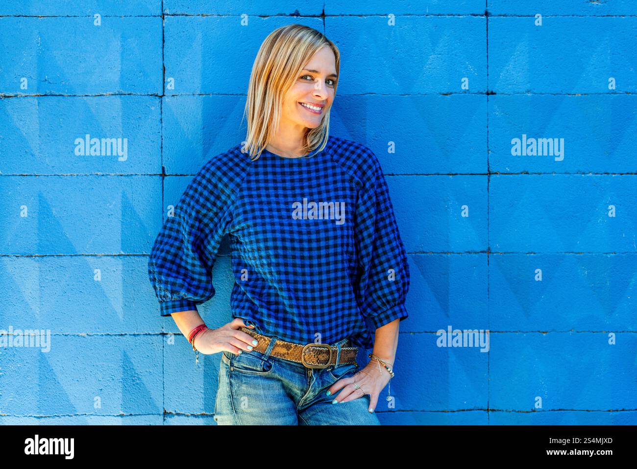 Una donna allegra con una camicia blu a scacchi posa casualmente con la mano sul fianco contro una parete blu vivace e testurizzata, guardando la fotocamera. Foto Stock