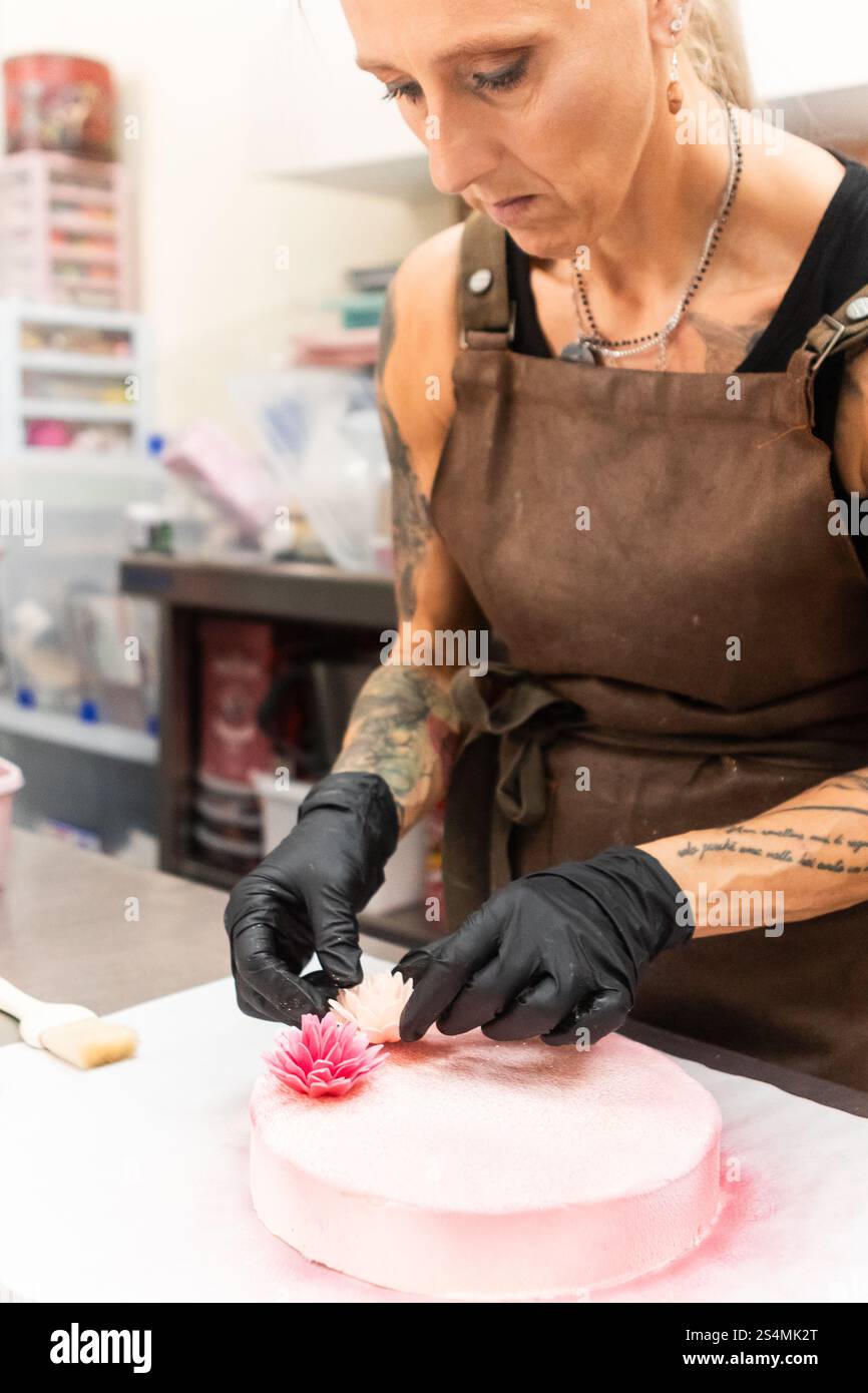 Un abile artigiano che indossa un grembiule e guanti aggiunge delicate decorazioni floreali a una pasticceria rosa, mettendo in mostra la meticolosa arte della panificazione italiana Foto Stock