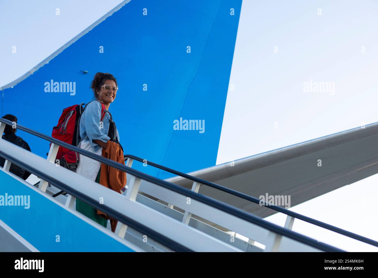 Un viaggiatore allegro sale le scale dell'aereo con uno zaino rosso contro un cielo blu brillante, la scena cattura l'emozione e l'attesa di una nuova Foto Stock