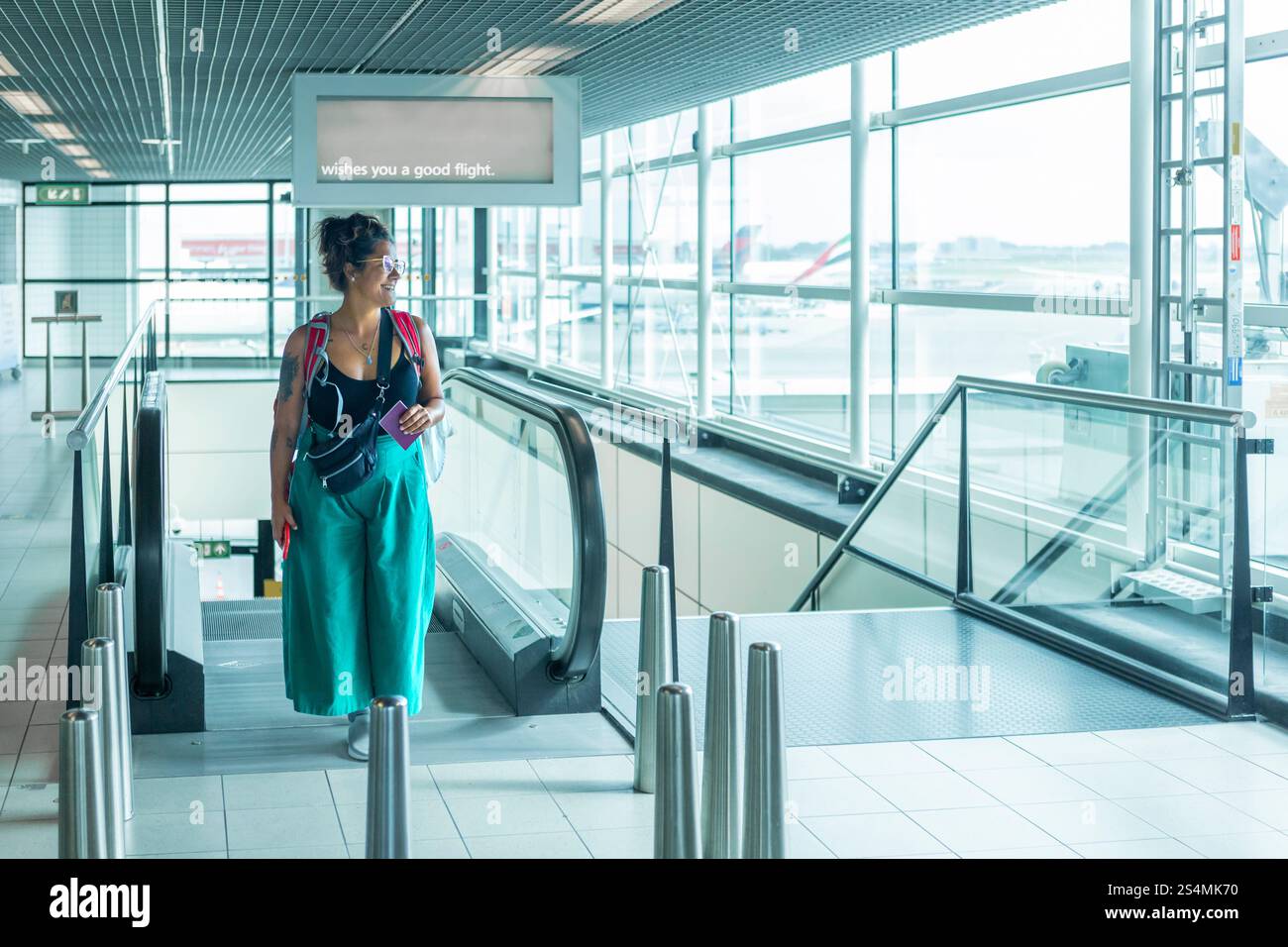 Un viaggiatore si trova su una scala mobile in un aeroporto luminoso, con in mano Un passaporto, Un cartello sopra vi augura un volo sicuro le pareti di vetro rivelano gli aerei all'esterno Foto Stock