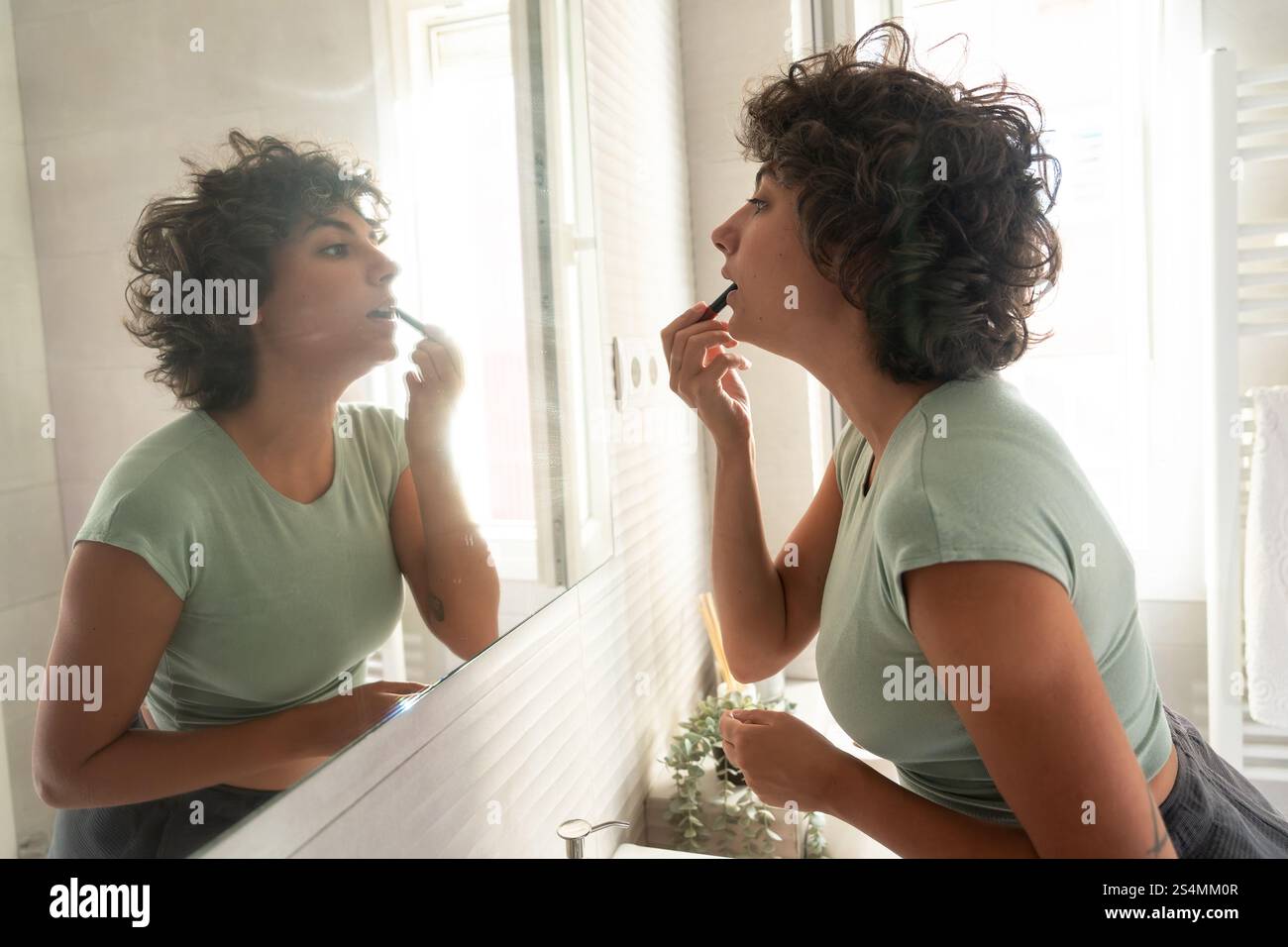 Una donna con i capelli ricci e la parte superiore verde applica il rossetto mentre guarda allo specchio di un bagno luminoso e illuminato dal sole, catturando un momento di bellezza mattutina Foto Stock