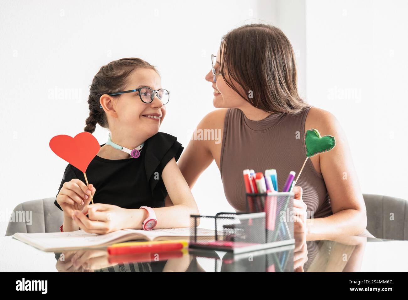 Ragazza disabile con tracheostomia, seduta con una sorella che la sostiene. Sono impegnati in un'interazione sentita, mostrando cuori di carta che sym Foto Stock