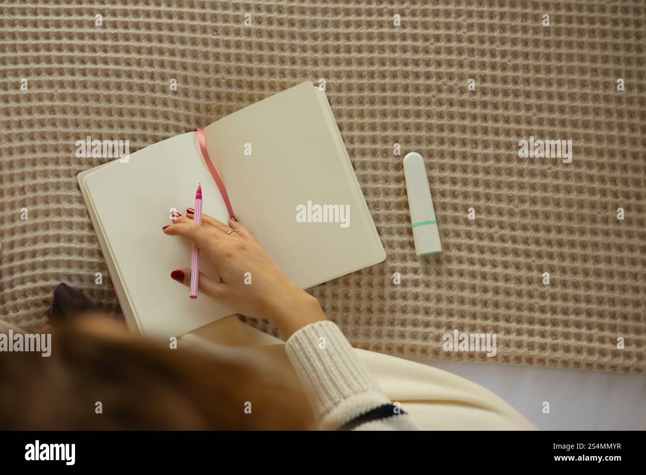 Una persona tiene una penna rosa sopra un quaderno aperto con un pennarello, posizionato su una coperta beige testurizzata, creando un'atmosfera accogliente ideale per il diario e. Foto Stock