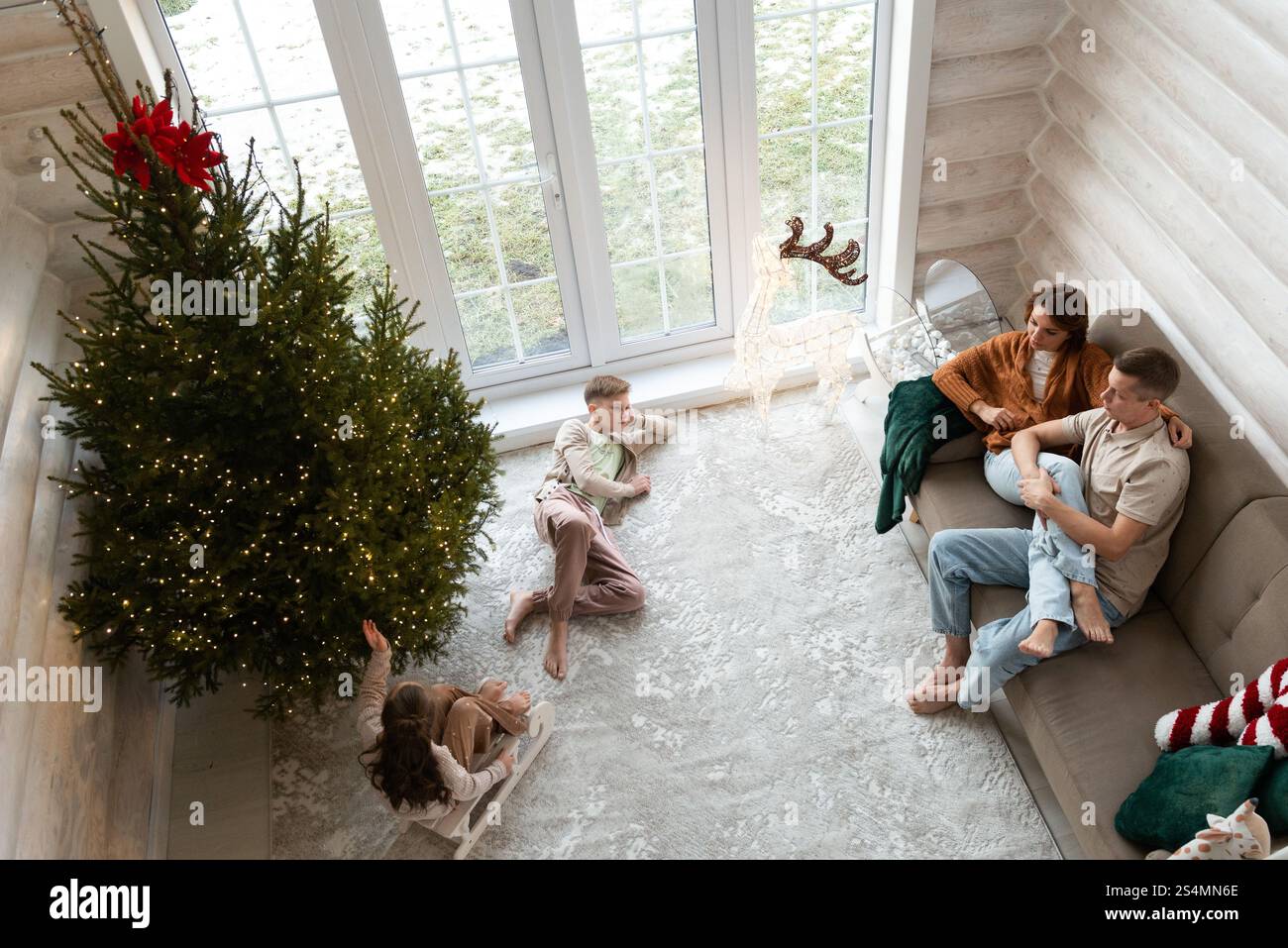 Una famiglia celebra il Natale in una camera accogliente adornata da un grande albero di Natale illuminato. Una coppia si siede su un divano due bambini giocano nelle vicinanze; una ragazza raggiunge Foto Stock