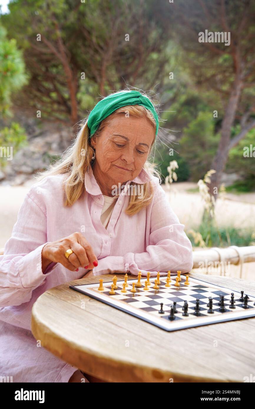 Una donna anziana gioca a scacchi all'aperto, profondamente concentrata sulle sue mosse strategiche. L'ambiente naturale e sereno aggiunge uno sfondo tranquillo a questo intellettuale pu Foto Stock