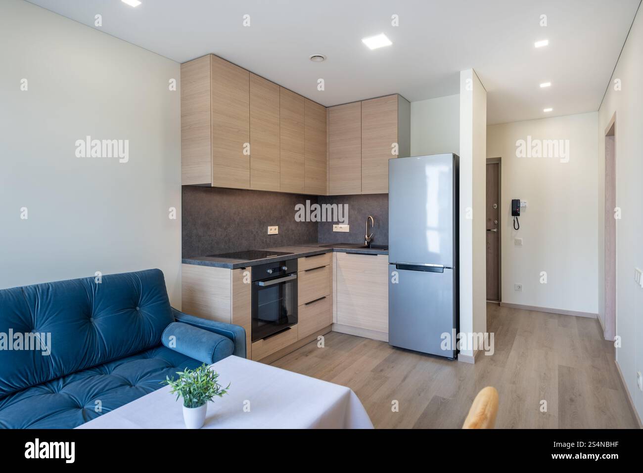 Cucina moderna e minimalista con armadi in legno, frigorifero argentato, accogliente divano soggiorno in un appartamento luminoso. Cucina minimalista e soggiorno con colazione continentale Foto Stock