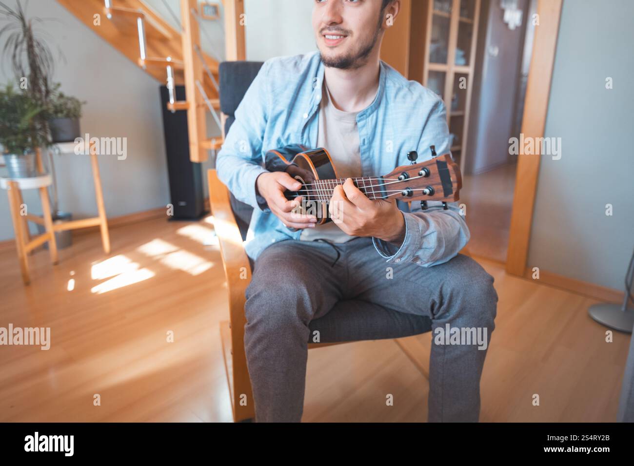 Un uomo allegro siede in una stanza luminosa, a suonare un ukulele nero. La sua camicia azzurra e la sua postura casual riflettono un momento musicale rilassato e gioioso Foto Stock