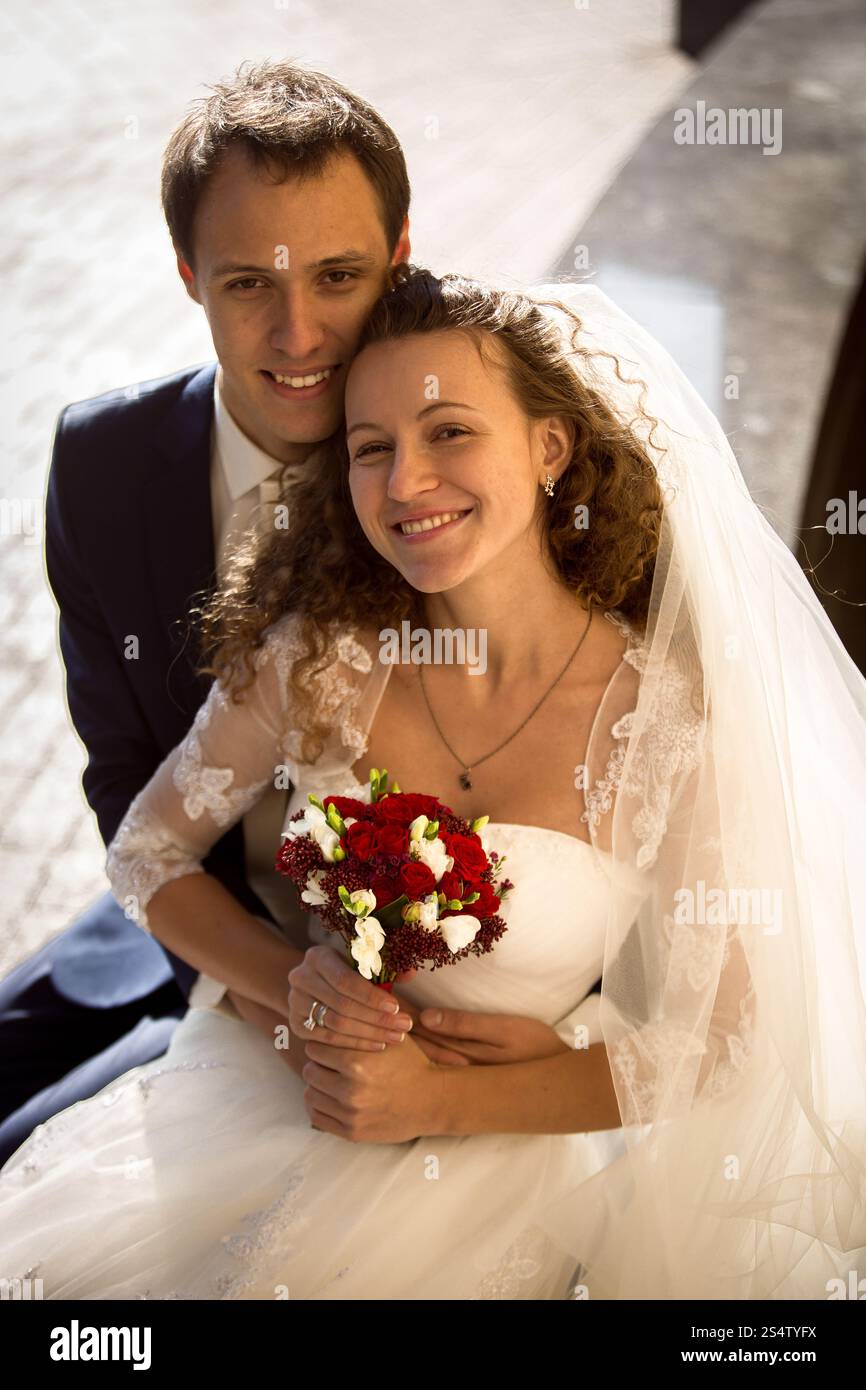 Bellissimo giovane sposo che abbraccia la sposa sorridente da dietro per strada Foto Stock