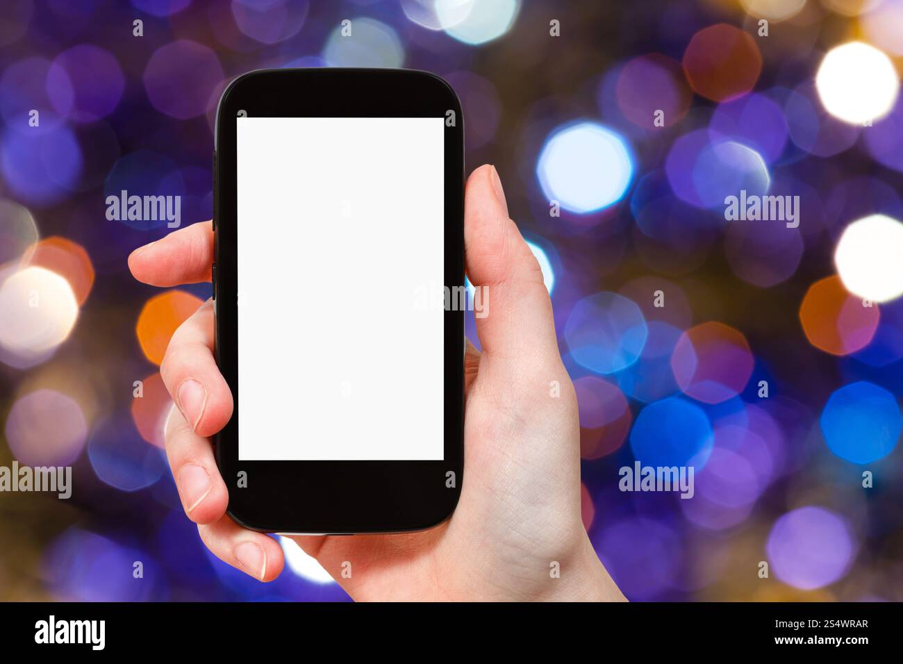 Concetto di festa di natale - mano con smartphone con schermo ritagliato sullo sfondo da luci natalizie blu scuro e viola scintillanti bokeh di Foto Stock