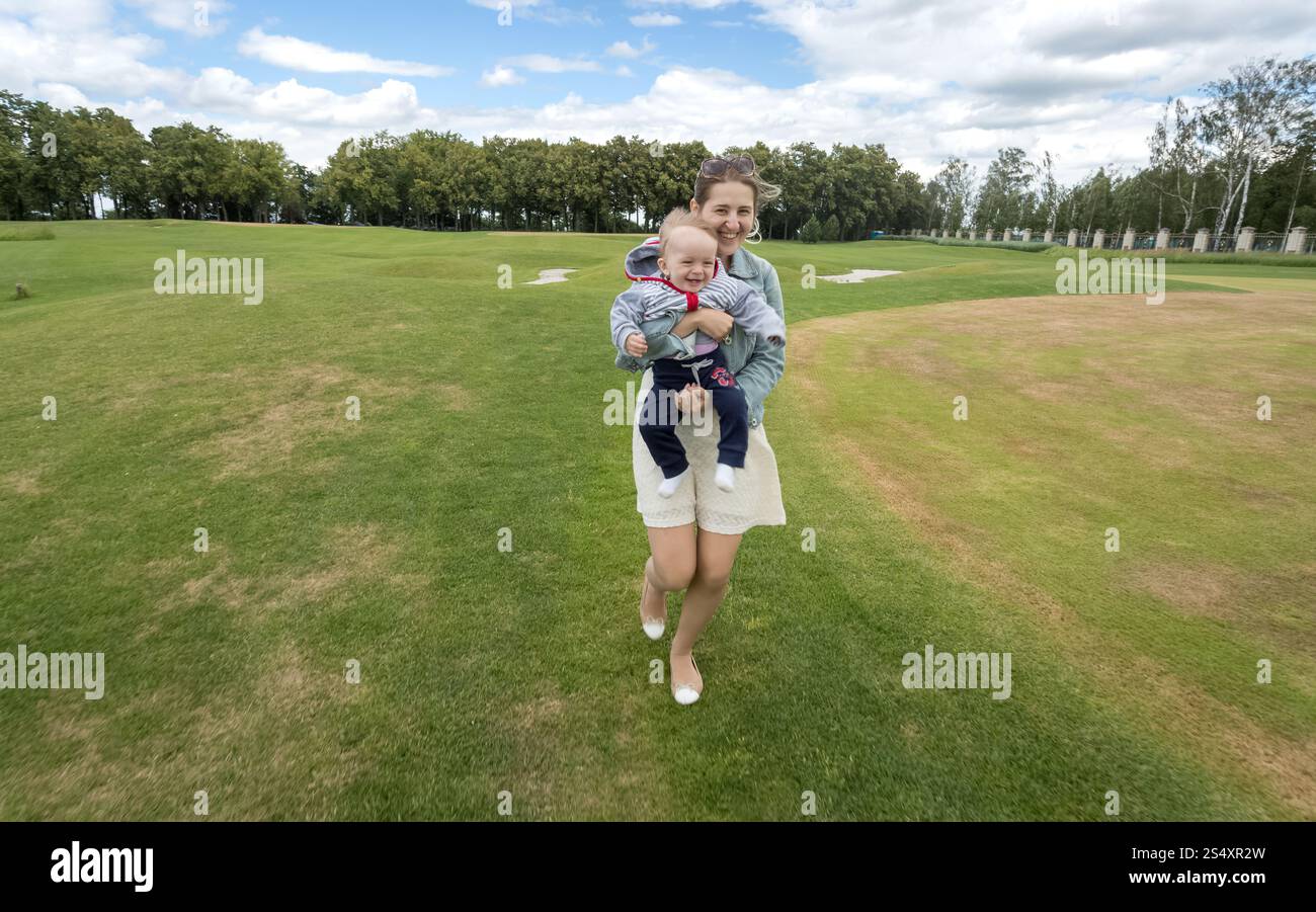 Bella madre allegra mentre tiene il suo bambino e in esecuzione su erba a park Foto Stock