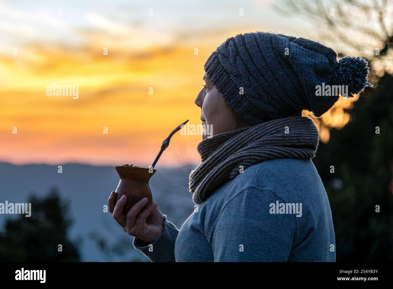 Donna in maglia con cappello e sciarpa che beve una yerba mate gourd al tramonto, con un paesaggio rurale panoramico sullo sfondo Foto Stock