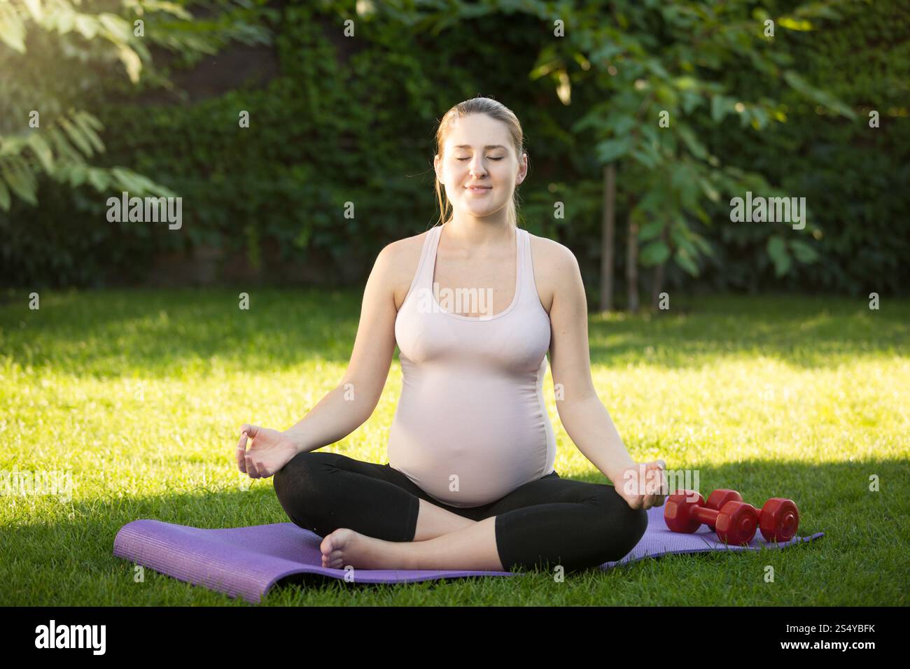 Bella donna incinta sorridente seduta in posa di loto sull'erba del parco Foto Stock