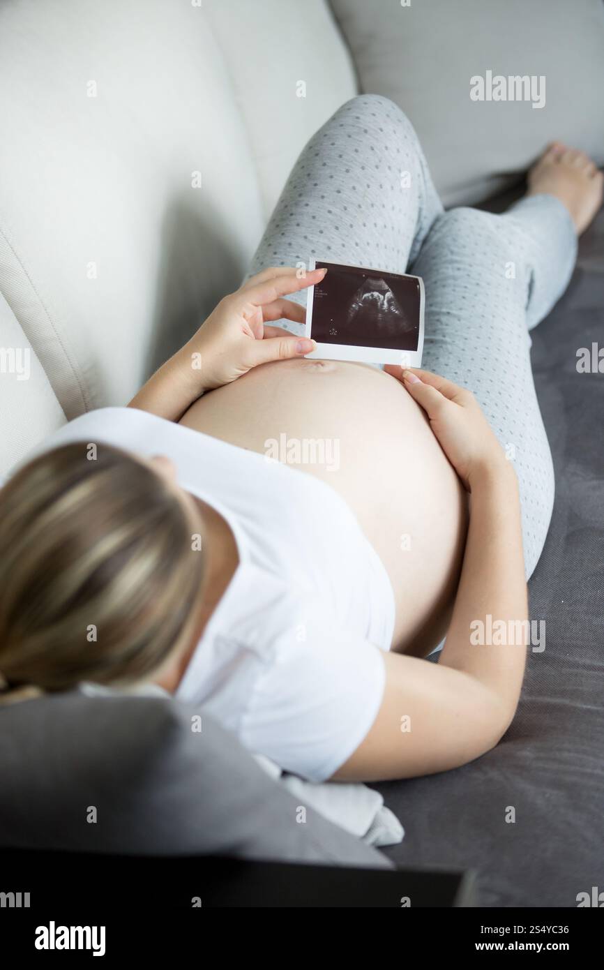 Foto ravvicinata di una giovane donna incinta che si rilassa sul divano e guarda la scansione del bambino a ultrasuoni Foto Stock