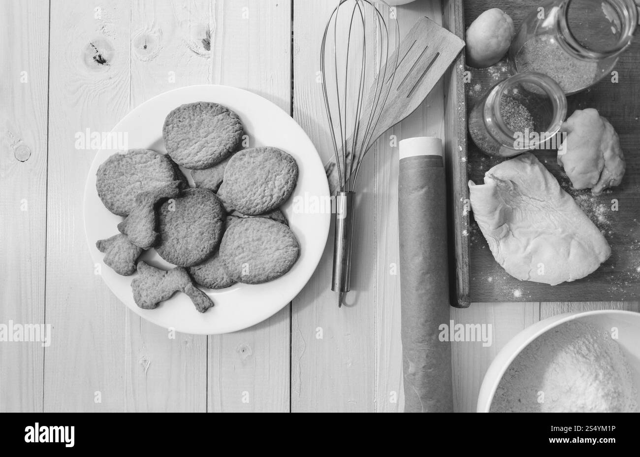 Foto in bianco e nero dell'impasto fresco steso su una scrivania di legno accanto agli utensili Foto Stock
