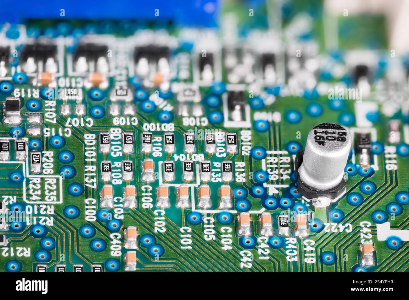 Condensatore elettrolitico bianco su scheda a circuito stampato con sfondo elettrotecnico sfocato. Primo piano di texture verde-blu con componenti elettronici. Foto Stock