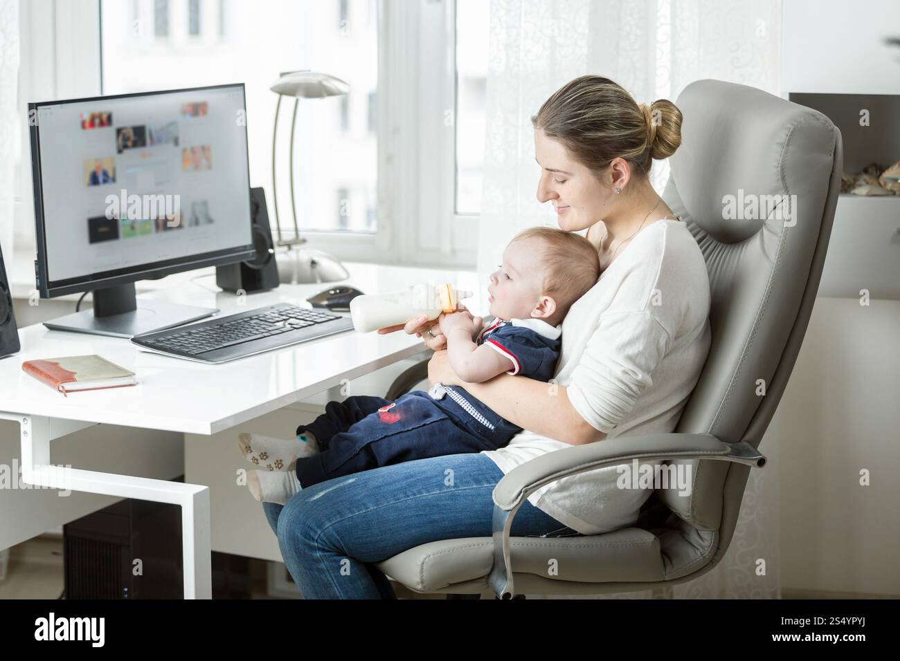 Mamma sorridente che lavora a casa e allatta il bimbo dal biberon Foto Stock